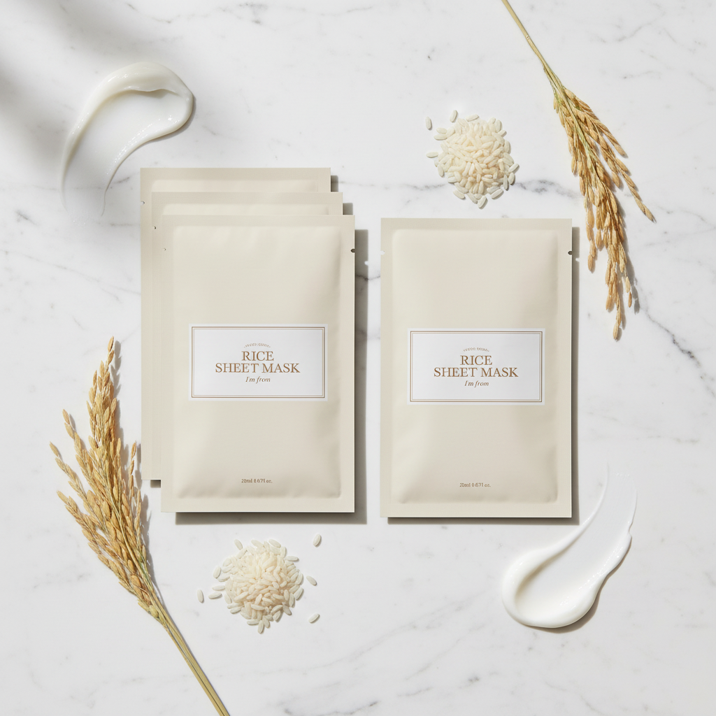 I'm From Rice Sheet Mask individual cream beige packet flat lay on white marble surface with raw Yeoju rice grains and rice stalks, premium K-beauty brightening sheet mask skincare editorial
